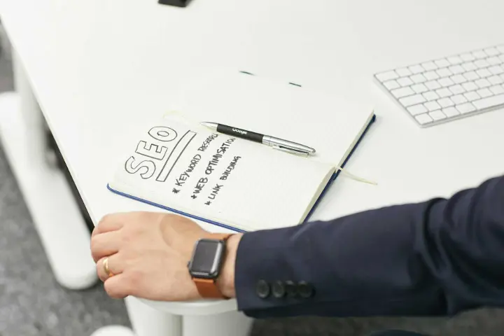 Caderno aberto com uma lista de verificação de SEO escrita à mão, incluindo pesquisa de palavras-chave, otimização da web e construção de links, ao lado da mão e do relógio de pulso de uma pessoa.