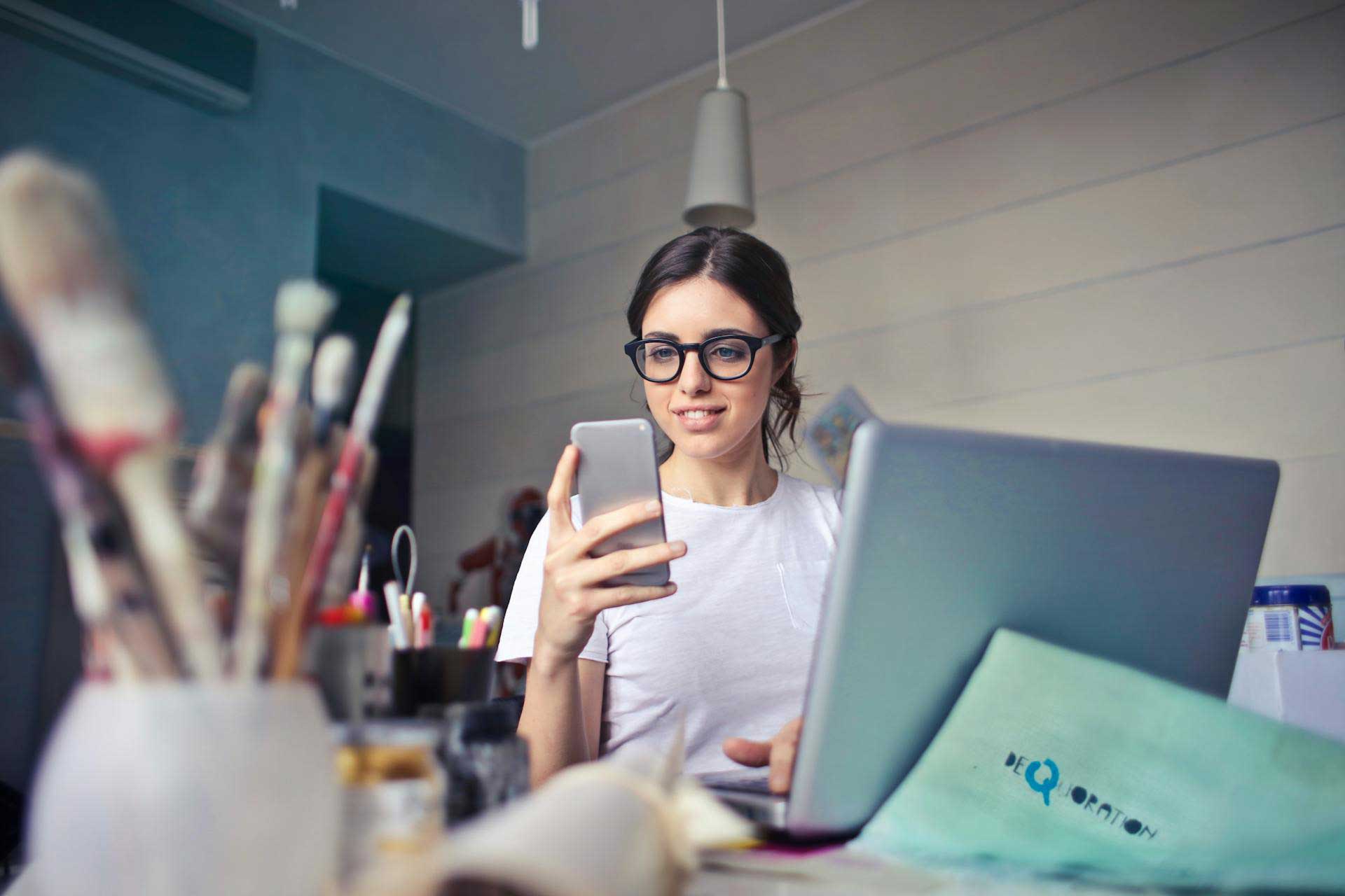 Mulher de óculos, olhando para um smartphone enquanto trabalha em um laptop em uma mesa abarrotada de materiais de arte.