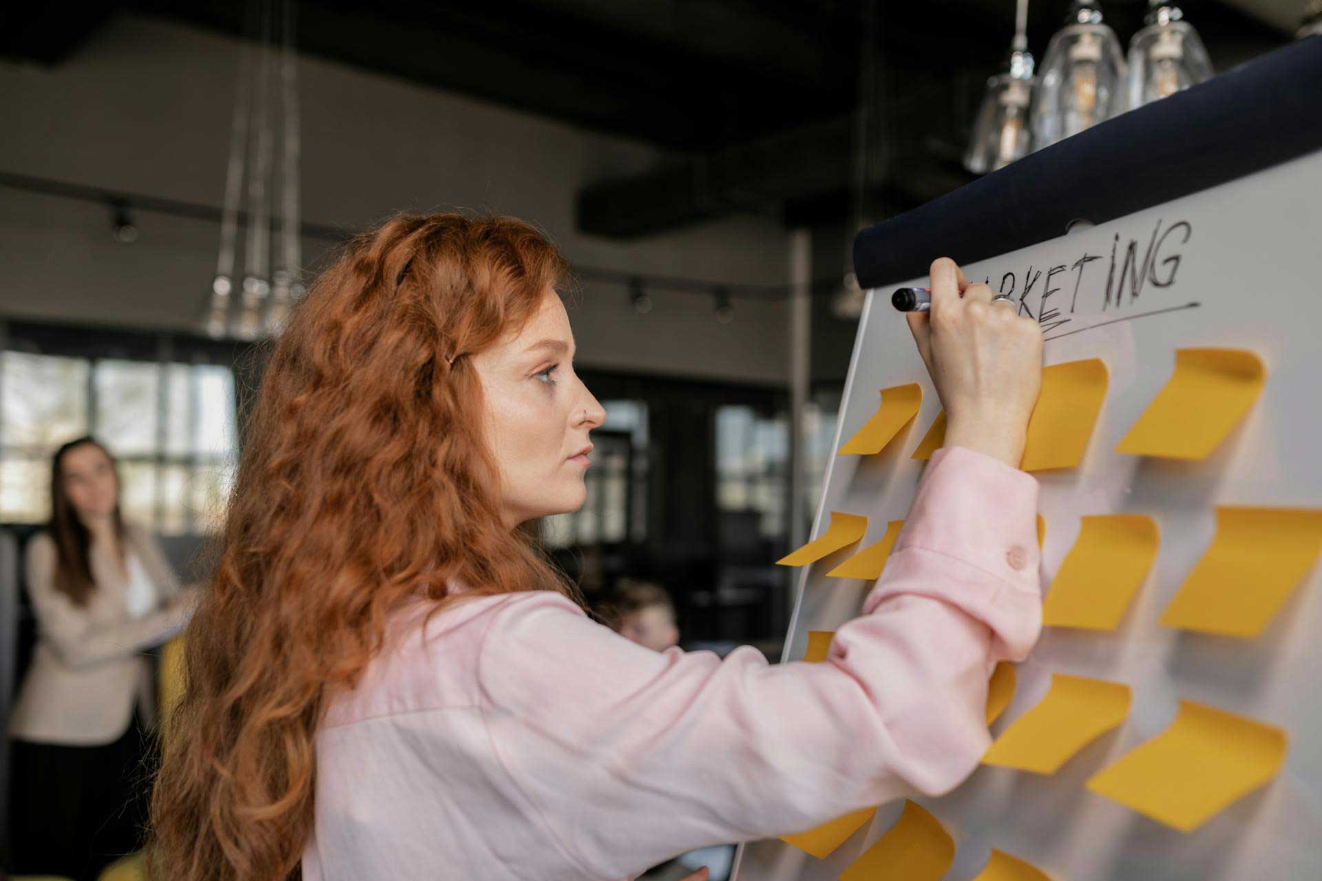 Mulher de cabelos ruivos escrevendo em um quadro branco com notas adesivas, focada em um plano de marketing.