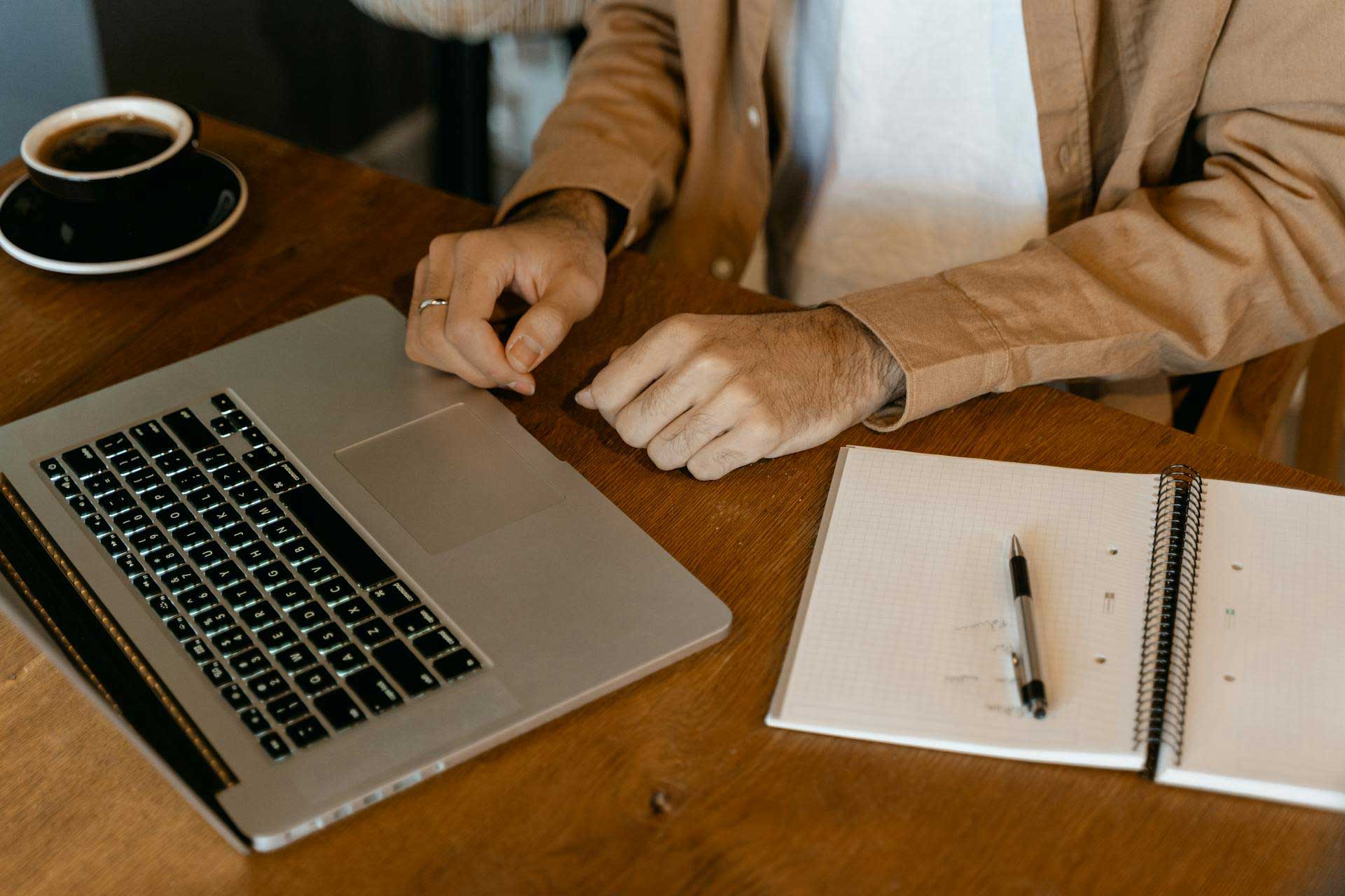 Pessoa trabalhando em uma mesa de madeira com um laptop, bloco de notas e caneta.