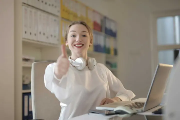 Mulher em um ambiente de escritório fazendo sinal de positivo, sorrindo e usando fones de ouvido.
