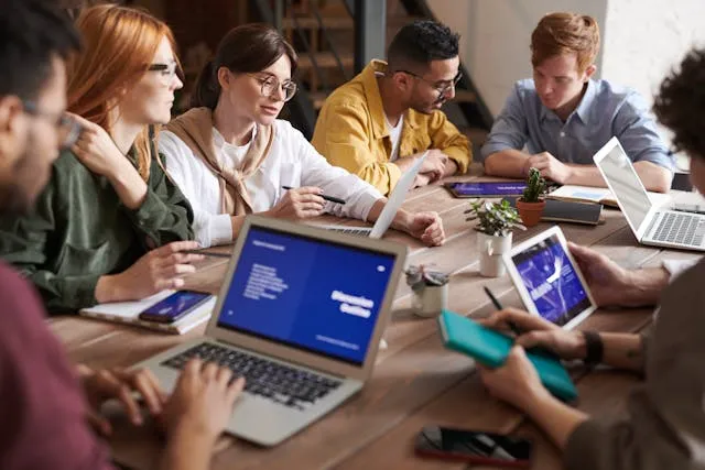 Grupo diversificado de pessoas colaborando em volta de uma mesa de madeira, trabalhando em laptops e tablets digitais.