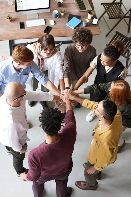 Grupo diversificado de pessoas com as mãos entrelaçadas formando um círculo, provavelmente em uma reunião colaborativa.