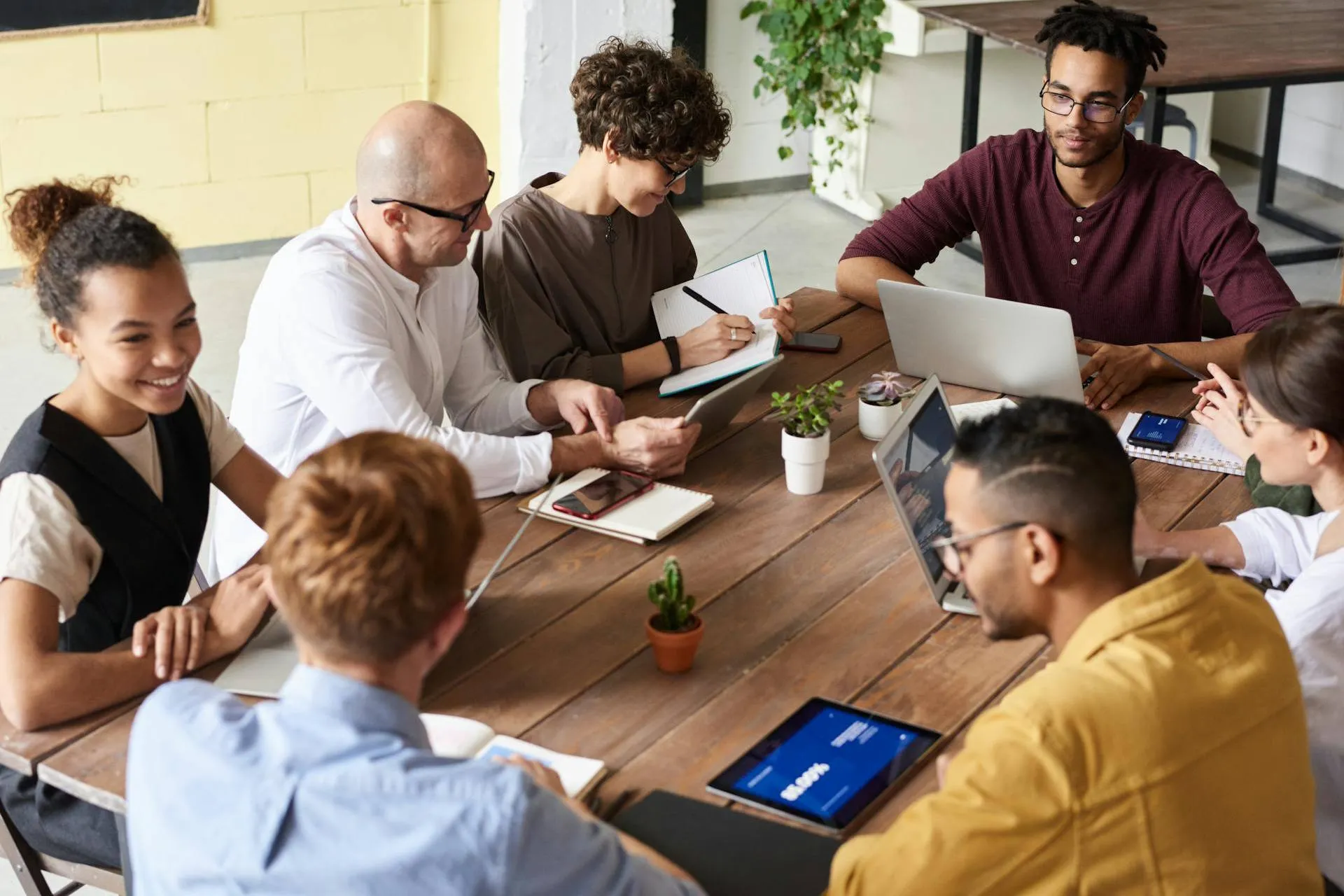 Grupo diversificado de pessoas em trajes casuais de negócios, sentadas ao redor de uma mesa de madeira, participando de uma reunião colaborativa com laptops e tablets.