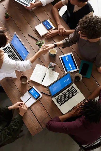 Grupo diversificado de pessoas sentadas ao redor de uma mesa de madeira, participando de uma reunião de negócios, com laptops, tablets e anotações visíveis, incluindo um aperto de mão.