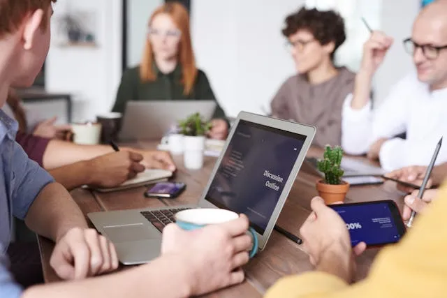 Grupo de pessoas em uma reunião, colaborando em laptops, tablets e dispositivos móveis, com um esboço da discussão visível em uma tela.