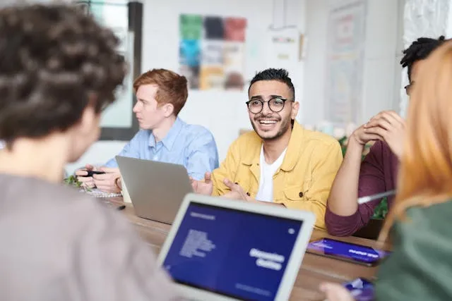 Grupo de pessoas em uma reunião, sentadas ao redor de uma mesa com laptops e um tablet, um homem de jaqueta amarela sorri e gesticula.