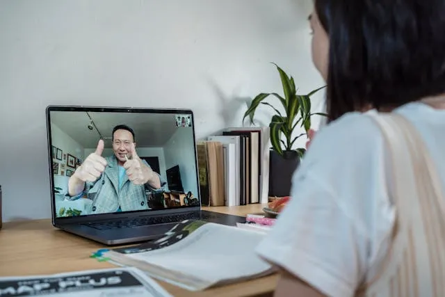 Pessoa fazendo sinal de positivo durante uma videochamada em um laptop, com livros e papéis sobre a mesa.